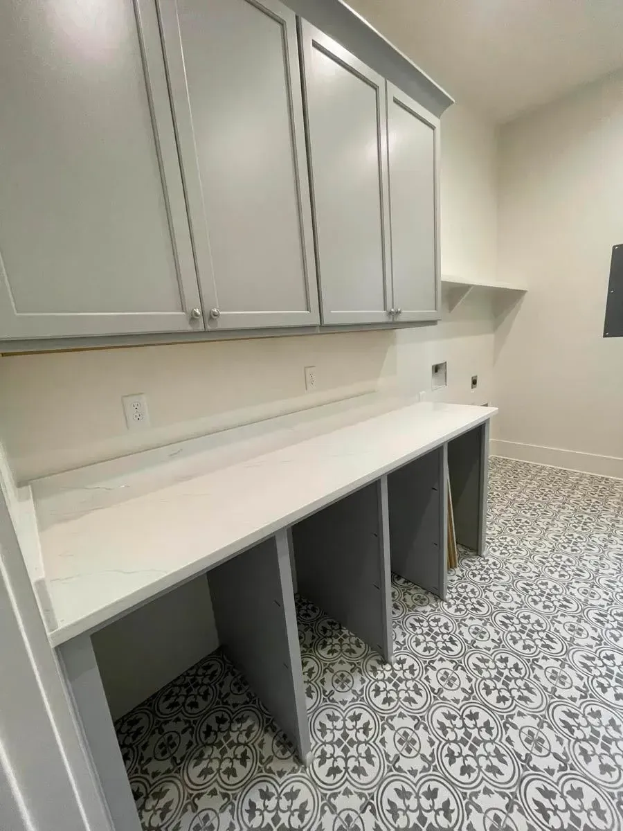 Laundry folding counter with open cubbies below and gray upper cabinets over patterned cement-look tile