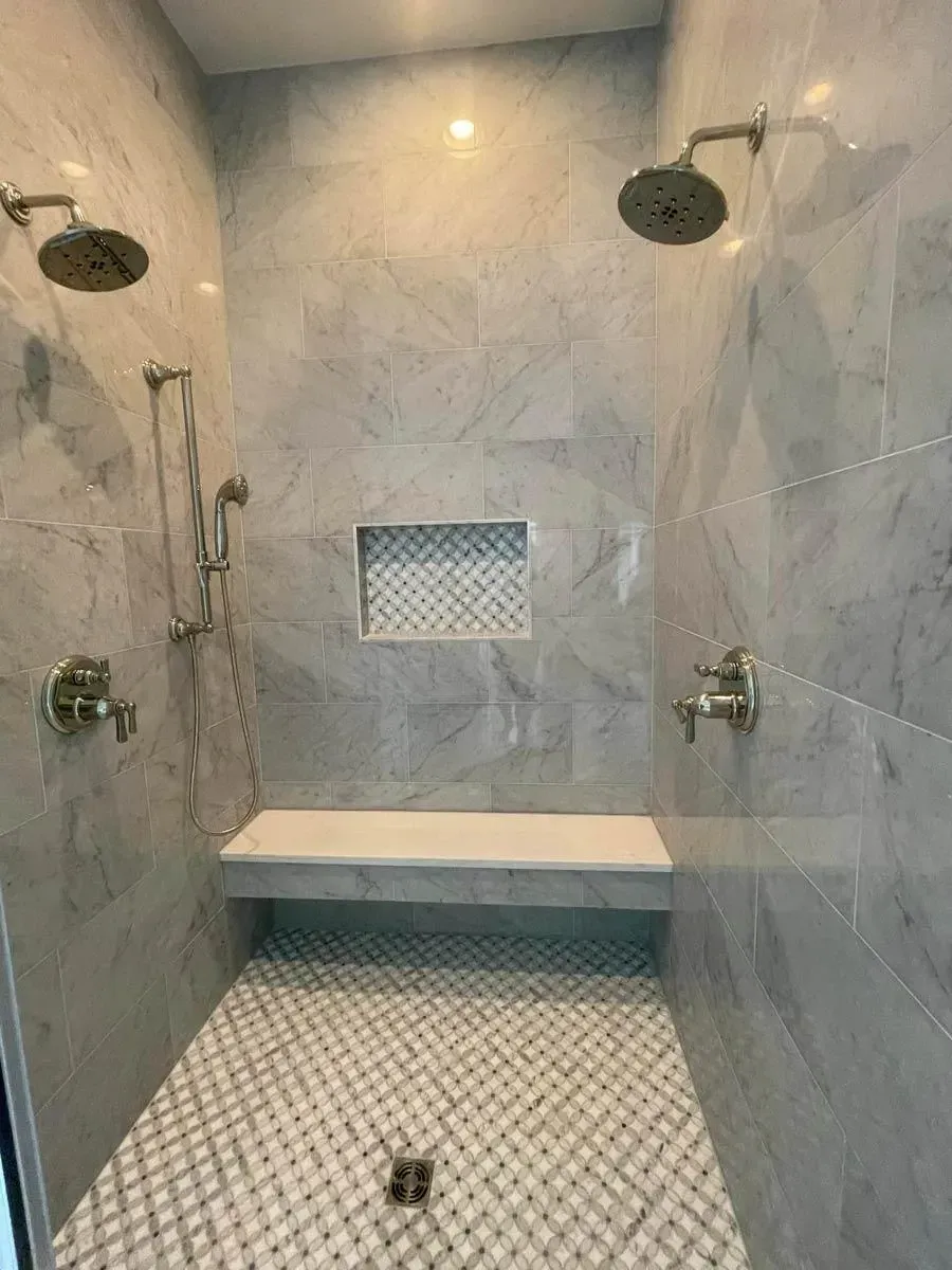 Primary shower clad in marble tile with two shower heads, a built-in bench, a niche and a mosaic floor
