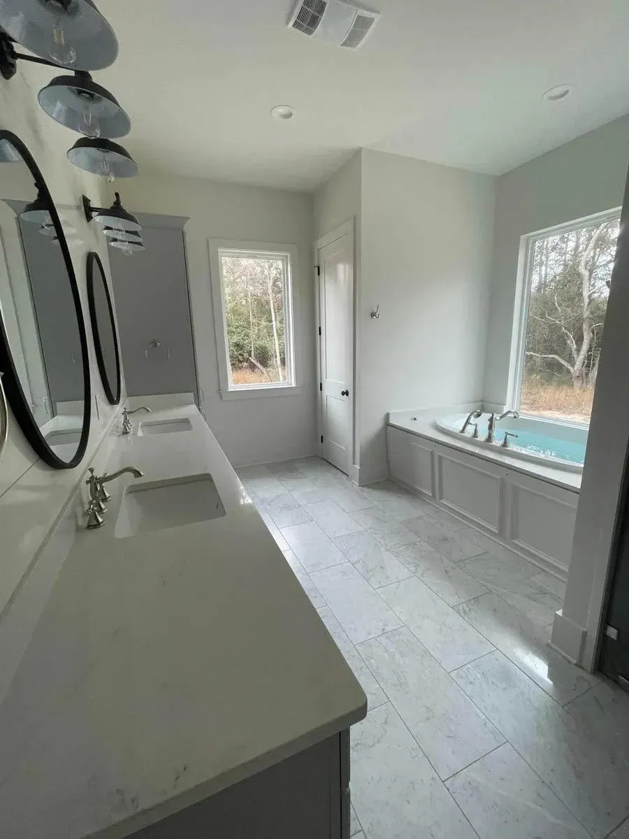 Primary bathroom with a gray double vanity and round black mirrors, marble-look floor tile and a soaking tub under the window