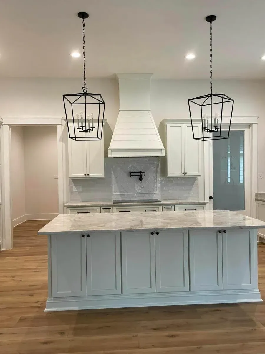 Kitchen island with white shaker cabinets and marble-look top under two black lantern pendants, with a white plaster range hood behind