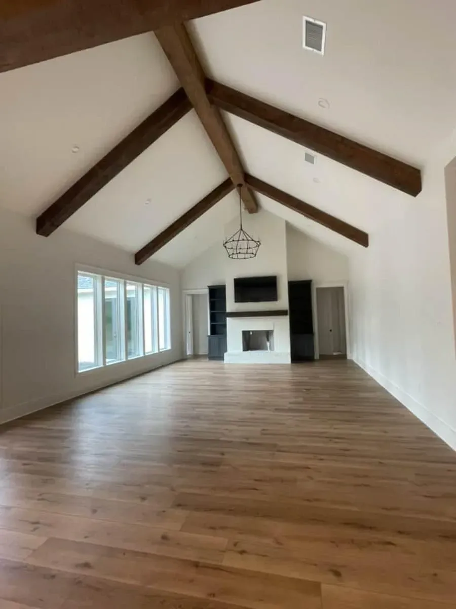 Great room with a cathedral ceiling and dark exposed beams, wide-plank oak floors, rear windows and the fireplace wall at the far end