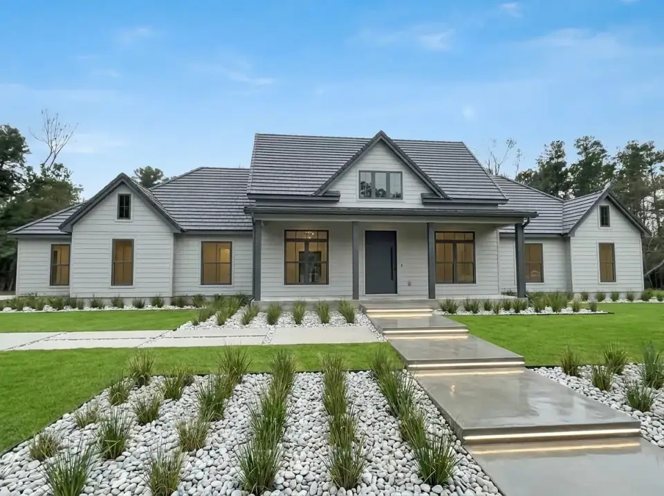 Plan 185028PHD in soft gray lap siding with a charcoal flat-tile roof, dark porch posts, a black front door and lit floating concrete steps through white river-stone beds planted with grasses