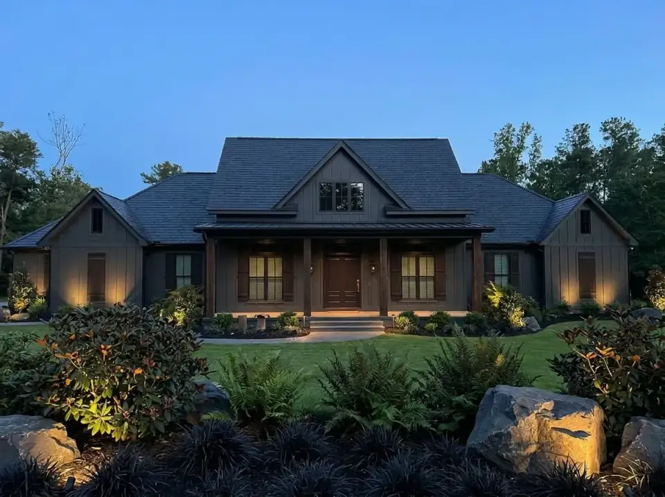 Plan 185028PHD at dusk in charcoal brown board-and-batten with a dark shingle roof, a metal porch roof, a wood front door and shutters, landscape uplighting on the gables, boulders and ferns